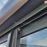 Szczegółowe zbliżenie na górny punkt styku panelu przesuwnego z systemową prowadnicą sufitową w rozwiązaniu ramowym Sotro. Aluminiowy profil skrzydła w kolorze ciemnego, matowego antracytu jest wyposażony w białą uszczelkę ślizgową o przekroju litery U, która stabilizuje panel na szynie, umożliwiając jego płynny ruch. Wzdłuż pionowej krawędzi profilu zamontowana jest zintegrowana uszczelka szczotkowa w kolorze szarym, która zapewnia szczelność konstrukcji i chroni wnętrze zabudowy przed czynnikami zewnętrznymi. Nad panelem widoczna jest masywna, obła osłona prowadnicy górnej, a w tle przezroczysta tafle szkła hartowanego odbija jasne, lekko zachmurzone niebo.