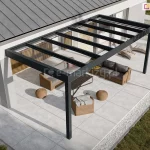 Wizualizacja przedstawia nowoczesną realizację zadaszenia tarasu Spekta Glass, zintegrowaną z bryłą minimalistycznego budynku jednorodzinnego o jasnej elewacji. Konstrukcja nośna została wykonana z profili aluminiowych w kolorze antracytowym, które tworzą ramę dachu opartą z jednej strony na ścianie budynku, a z drugiej na dwóch pionowych słupach. Dach konstrukcji jest w całości przeszklony i podzielony na regularne sekcje, co zapewnia pełną przezierność i ochronę przed warunkami atmosferycznymi. Pod zadaszeniem zaaranżowano strefę relaksu z meblami z technorattanu w odcieniu naturalnego brązu, obejmującą sofę, fotel oraz niskie pufy i stolik kawowy. Podłoże tarasu oraz opaskę wokół budynku stanowią wielkoformatowe płyty tarasowe w kolorze jasnoszarym, które są oddzielone od trawnika pasem białego gresu. Architektura ogrodu obejmuje również ozdobne trawy w białych donicach oraz nasadzenia wzdłuż elewacji, co tworzy spójną i estetyczną przestrzeń wypoczynkową.