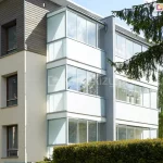 Zdjęcie przedstawia nowoczesną zabudowę balkonów w bloku mieszkalnym, wykonaną przy użyciu systemu szklanych drzwi przesuwnych Sotro. Trzykondygnacyjna sekcja budynku posiada balkony całkowicie zamknięte szklanymi panelami, które są osadzone w białych profilach aluminiowych, tworząc estetyczne i jednolite przeszklenia od balustrady aż po sufit każdego poziomu. Górne części zabudowy na każdym piętrze składają się z szerokich, przesuwnych tafli szkła hartowanego, natomiast dolne partie pod linią balustrad wypełnione są mlecznym lub nieprzezroczystym szkłem, co zapewnia prywatność mieszkańcom. Konstrukcja jest zintegrowana z jasną elewacją budynku oraz wstawkami z ciemnego drewna na najwyższym piętrze, a w szklanych panelach odbija się błękit nieba i okoliczne drzewa. Widok od strony ogrodu z gęstym żywopłotem podkreśla funkcjonalność systemu w tworzeniu dodatkowej, chronionej przed hałasem i pogodą przestrzeni użytkowej w budownictwie wielorodzinnym.