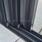 Zbliżenie na dolny mechanizm jezdny trzech skrzydeł drzwiowych systemu Sotro, osadzonych w wielotorowej prowadnicy dolnej. Panele są ułożone kaskadowo, co ukazuje sposób ich nachodzenia na siebie w pozycji otwartej. Każde skrzydło ujęte jest w pionowy profil aluminiowy o matowym wykończeniu w kolorze antracytowym, który w dolnej części posiada białą uszczelkę ślizgową w kształcie odwróconej litery U, obejmującą tor prowadnicy. Prowadnica o niskim profilu jest zamontowana na styku dwóch rodzajów nawierzchni: jasnych płytek tarasowych po lewej stronie oraz gładkiej wylewki betonowej po stronie prawej. Na bocznej płaszczyźnie pierwszego profilu widoczna jest również okrągła zaślepka otworu montażowego, a precyzyjne spasowanie elementów podkreśla techniczną jakość systemu przesuwnego.