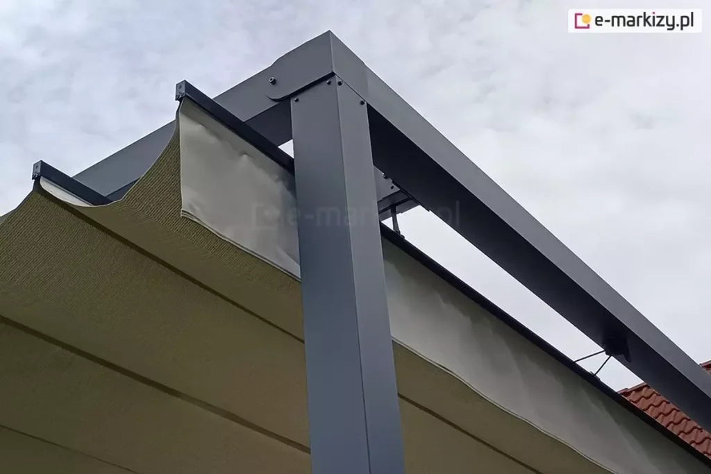 Zbliżenie na narożnik konstrukcji nowoczesnej pergoli tarasowej wykonanej z profili aluminiowych w kolorze antracytowym (ciemnoszarym). Zdjęcie prezentuje detal połączenia pionowego słupa wsporczego o kwadratowym przekroju z poziomymi belkami ramy. Pod ramą widoczne jest poszycie z tkaniny technicznej w kolorze kremowym/beżowym, które jest ułożone w charakterystyczne fałdy systemu rzymskiego. Tkanina jest zamocowana na poziomych profilach usztywniających (poprzeczkach), które poruszają się wewnątrz prowadnic zintegrowanych z bocznymi belkami konstrukcji. Widoczne są elementy montażowe, takie jak śruby imbusowe oraz czarne zaślepki końcowe profili tkaniny. W tle znajduje się jasne, zachmurzone niebo oraz fragment ceramicznej dachówki w kolorze ceglastym.