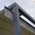Zbliżenie na narożnik konstrukcji nowoczesnej pergoli tarasowej wykonanej z profili aluminiowych w kolorze antracytowym (ciemnoszarym). Zdjęcie prezentuje detal połączenia pionowego słupa wsporczego o kwadratowym przekroju z poziomymi belkami ramy. Pod ramą widoczne jest poszycie z tkaniny technicznej w kolorze kremowym/beżowym, które jest ułożone w charakterystyczne fałdy systemu rzymskiego. Tkanina jest zamocowana na poziomych profilach usztywniających (poprzeczkach), które poruszają się wewnątrz prowadnic zintegrowanych z bocznymi belkami konstrukcji. Widoczne są elementy montażowe, takie jak śruby imbusowe oraz czarne zaślepki końcowe profili tkaniny. W tle znajduje się jasne, zachmurzone niebo oraz fragment ceramicznej dachówki w kolorze ceglastym.