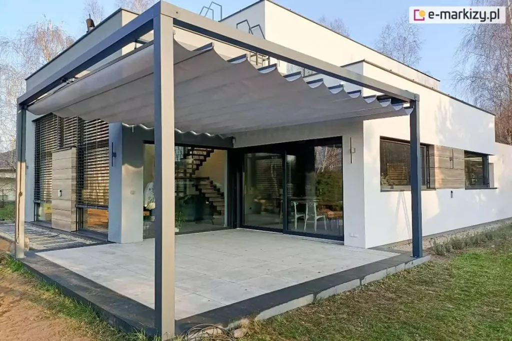 Nowoczesna pergola tarasowa wolnostojąca Tako o grafitowej konstrukcji aluminiowej, która osłania duży, jasny taras. Zadaszenie wykonane jest z popielatej tkaniny, która układa się w eleganckie, faliste segmenty. Pergola Tako doskonale komponuje się z minimalistyczną architekturą białego, piętrowego domu o prostej bryle. Przez duże przeszklenia budynku widać nowoczesne wnętrze z ażurowymi schodami. Taras wyłożony jest dużymi, jasnoszarymi płytami, a wokół rozciąga się zadbany trawnik oraz niskie nasadzenia z lawendy. Całość sfotografowana w świetle dziennym, co podkreśla czyste linie konstrukcji i nowoczesny charakter posiadłości.