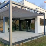Nowoczesna pergola tarasowa wolnostojąca Tako o grafitowej konstrukcji aluminiowej, która osłania duży, jasny taras. Zadaszenie wykonane jest z popielatej tkaniny, która układa się w eleganckie, faliste segmenty. Pergola Tako doskonale komponuje się z minimalistyczną architekturą białego, piętrowego domu o prostej bryle. Przez duże przeszklenia budynku widać nowoczesne wnętrze z ażurowymi schodami. Taras wyłożony jest dużymi, jasnoszarymi płytami, a wokół rozciąga się zadbany trawnik oraz niskie nasadzenia z lawendy. Całość sfotografowana w świetle dziennym, co podkreśla czyste linie konstrukcji i nowoczesny charakter posiadłości.