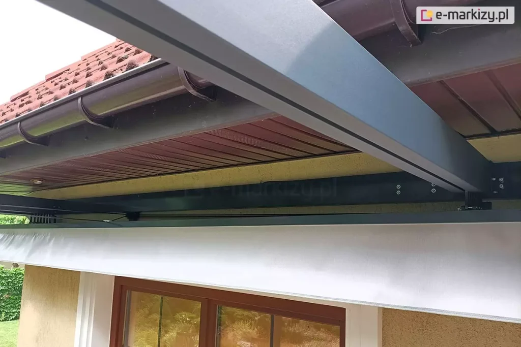 Zbliżenie na system montażu przyściennego pergoli tarasowej w kolorze antracytowym. Zdjęcie przedstawia punkt styku konstrukcji z elewacją budynku, tuż pod podbitką dachową. Główna belka nośna pergoli jest przymocowana do ściany za pomocą solidnych uchwytów i śrub montażowych. Nad belką widoczny jest fragment brązowej rynny oraz drewnianej podbitki. Poniżej profilu nośnego widoczny jest dach rzymski z belkami przeciwwietrznymi, z których wysunięty jest fragment jasnej, białej tkaniny technicznej. Konstrukcja jest precyzyjnie dopasowana do nadproża okiennego – w dolnej części kadru widać fragment ramy okna tarasowego w kolorze brązowym (drewnopodobnym). Detal podkreśla estetyczne wykończenie instalacji oraz stabilność mocowania elementów mechanicznych do struktury budynku.