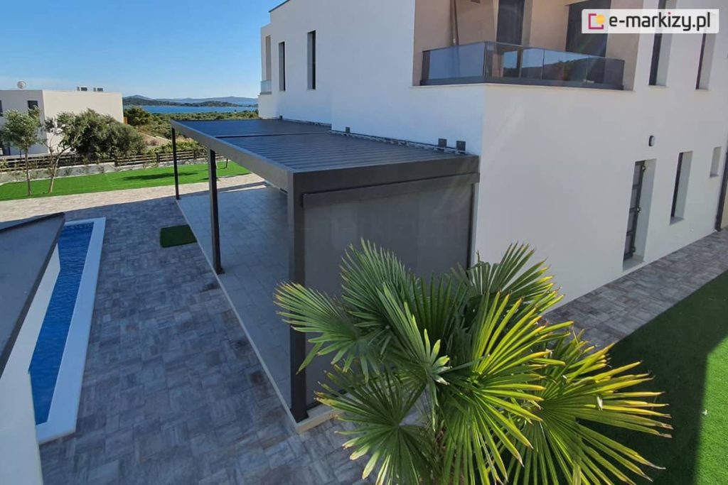 Pergola przyścienna z lamelami ustawionymi prostopadle do ściany, dzięki dwóm modułom uzyskano spójne zadaszenie dla tarasu o nietypowym kształcie