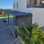 Pergola przyścienna z lamelami ustawionymi prostopadle do ściany, dzięki dwóm modułom uzyskano spójne zadaszenie dla tarasu o nietypowym kształcie