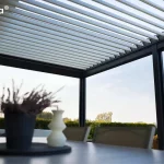 Widok z bliska na spód zadaszenia nowoczesnej pergoli essential Tarasola w ciemnym kolorze, prezentujący otwarty dach lamelowy. Białe, aluminiowe lamele są ustawione pod kątem, co pozwala na regulację dostępu światła słonecznego oraz naturalną wentylację przestrzeni tarasowej. Konstrukcja dachu opiera się na solidnych, czarnych profilach bocznych, które kontrastują z jasnym kolorem piór dachu. Na pierwszym planie, w rozmyciu, widoczny jest fragment stołu z dekoracyjną rośliną w doniczce oraz świecą, co podkreśla salonowy charakter aranżacji. W tle, pod konstrukcją, rozpościera się widok na zielony ogród z żywopłotem oraz jasne, pogodne niebo, ukazując funkcjonalność pergoli bioklimatycznej w słoneczny dzień.