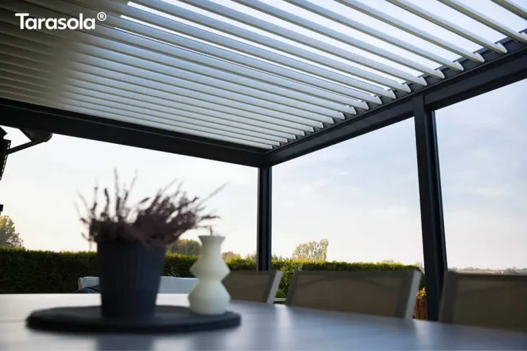 Widok z bliska na spód zadaszenia nowoczesnej pergoli essential Tarasola w ciemnym kolorze, prezentujący otwarty dach lamelowy. Białe, aluminiowe lamele są ustawione pod kątem, co pozwala na regulację dostępu światła słonecznego oraz naturalną wentylację przestrzeni tarasowej. Konstrukcja dachu opiera się na solidnych, czarnych profilach bocznych, które kontrastują z jasnym kolorem piór dachu. Na pierwszym planie, w rozmyciu, widoczny jest fragment stołu z dekoracyjną rośliną w doniczce oraz świecą, co podkreśla salonowy charakter aranżacji. W tle, pod konstrukcją, rozpościera się widok na zielony ogród z żywopłotem oraz jasne, pogodne niebo, ukazując funkcjonalność pergoli bioklimatycznej w słoneczny dzień.