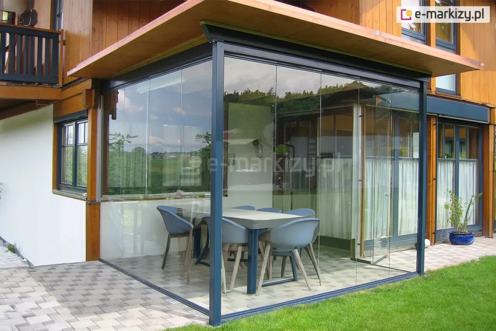 Zabudowany narożny taras przylegający do drewniano-murowanego domu jednorodzinnego. Konstrukcja wykonana z ciemnych profili aluminiowych oraz przezroczystych, bezramowych paneli szklanych Miros, które tworzą lekką, nowoczesną osłonę przestrzeni jadalnej. Wewnątrz znajduje się stół prostokątny oraz sześć nowoczesnych krzeseł w odcieniach szarości i błękitu. Podłoże wyłożone płytkami w odcieniu jasnego beżu. Po lewej stronie fragment elewacji z drewnianymi detalami i czarnymi oknami, po prawej – ogród z trawnikiem i donicą. W szybach odbija się krajobraz z zielonymi polami i drzewami.