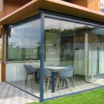 Zabudowany narożny taras przylegający do drewniano-murowanego domu jednorodzinnego. Konstrukcja wykonana z ciemnych profili aluminiowych oraz przezroczystych, bezramowych paneli szklanych Miros, które tworzą lekką, nowoczesną osłonę przestrzeni jadalnej. Wewnątrz znajduje się stół prostokątny oraz sześć nowoczesnych krzeseł w odcieniach szarości i błękitu. Podłoże wyłożone płytkami w odcieniu jasnego beżu. Po lewej stronie fragment elewacji z drewnianymi detalami i czarnymi oknami, po prawej – ogród z trawnikiem i donicą. W szybach odbija się krajobraz z zielonymi polami i drzewami.