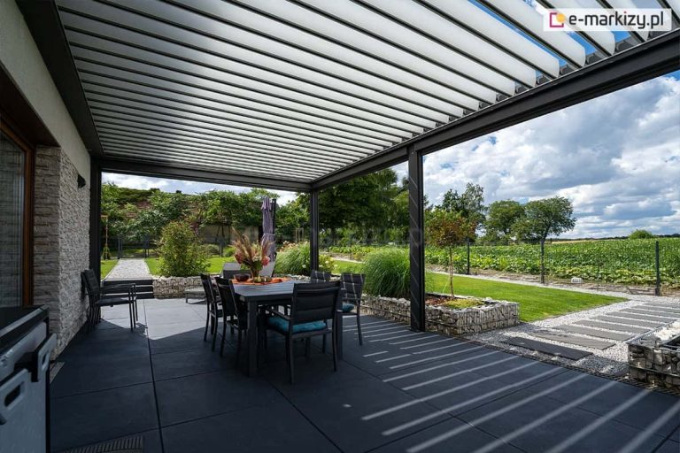 Wnętrze nowoczesnej pergoli bioklimatycznej SB400 o konstrukcji aluminiowej w kolorze antracytowym, zamontowanej na tarasie z ciemnych, wielkoformatowych płyt. Dach pergoli składa się z białych, poziomych lameli, które są częściowo otwarte, rzucając pasy cienia na podłoże. Pod zadaszeniem znajduje się zestaw mebli ogrodowych: ciemny stół z krzesłami wyposażonymi w niebieskie poduszki. Na stole stoi wazon z kwiatami i dekoracyjna misa. Po lewej stronie widoczna jest elewacja domu wyłożona jasnym, dekoracyjnym kamieniem. Taras graniczy z ogrodem, w którym znajdują się gabiony wypełnione jasnym kruszywem, ozdobne trawy, niewielkie drzewka oraz ścieżki z szarych płyt chodnikowych. W tle rozpościera się widok na zielone pole uprawne, drzewa oraz zachmurzone niebo.