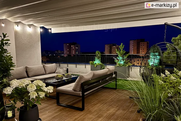 Zdjęcie prezentuje luksusową aranżację tarasu na dachu budynku wielorodzinnego, wyposażonego w system zadaszenia tkaninowego Supro. Jasna tkanina dachu rzymskiego jest całkowicie rozwinięta, tworząc nad całą strefą wypoczynku gładką płaszczyznę z delikatnymi, poziomymi przetłoczeniami w miejscach profili wspierających. Taras został urządzony w nowoczesnym stylu z wykorzystaniem mebli modułowych z ciemnymi stelażami i szarymi poduszkami, ustawionych wokół niskiego stolika kawowego. Nawierzchnia łączy naturalne deski kompozytowe w ciepłym odcieniu brązu z fragmentami sztucznej trawy oraz jasnym żwirem dekoracyjnym. Przestrzeń jest bogato obsadzona roślinnością w donicach, w tym białymi hortensjami i ozdobnymi trawami. Klimat wieczorny podkreśla nastrojowe oświetlenie w formie girlandy żarówkowej (typu outdoor) zawieszonej pod dachem oraz punktowe podświetlenie roślin w kolorze zielonym. W tle, za metalową balustradą, widoczna jest panorama miasta z oświetlonymi blokami mieszkalnymi pod nocnym niebem.