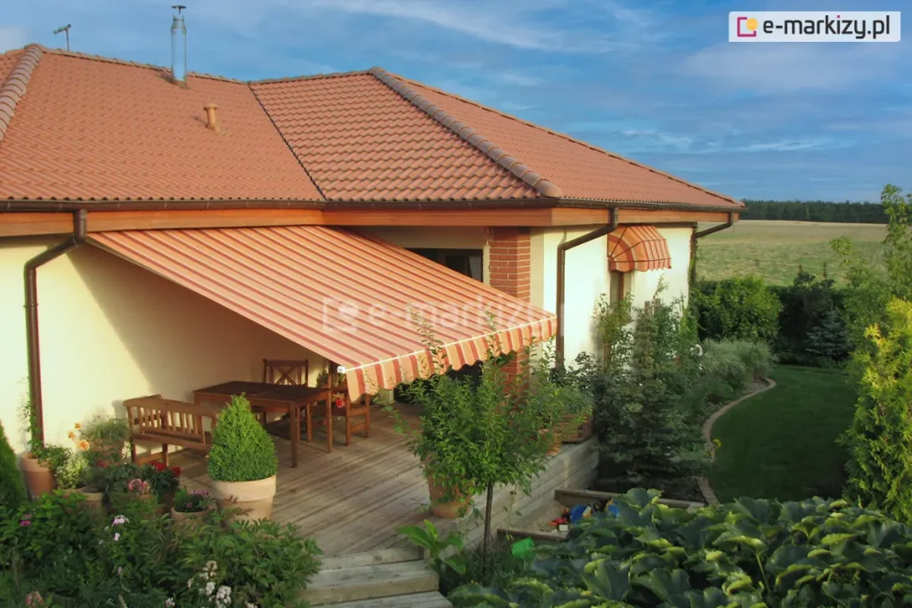 Zdjęcie prezentuje dom jednorodzinny o jasnej, kremowej elewacji z zainstalowaną markizą Mezzo mol, która osłania drewniany taras. Markiza charakteryzuje się poszyciem w ciepłej kolorystyce, składającym się z naprzemiennych pasów w odcieniach pomarańczu, czerwieni i beżu. Tkanina jest w pełni rozłożona pod kątem, wsparta na bocznych ramionach, a jej krawędź wykończona jest falistą falbaną z białą lamówką. Budynek posiada dach wielospadowy pokryty ceglastą dachówką ceramiczną, z wyraźnie zaznaczonymi brązowymi rynnami i podbitką dachową w kolorze jasnego drewna. Na tarasie pod markizą Mezzo mol znajduje się drewniany komplet mebli: prostokątny stół, krzesła z ażurowymi oparciami oraz ławka. Przestrzeń wokół tarasu jest bogato zagospodarowana roślinnością – widoczne są liczne krzewy ozdobne, iglaki oraz kwiaty w ceramicznych donicach. Po prawej stronie, nad mniejszym oknem, zamontowana jest dodatkowa, mniejsza markiza koszowa o identycznym wzorze materiału. W tle rozpościera się widok na zielony ogród z krętą ścieżką oraz daleki horyzont z linią lasu pod błękitnym, lekko zachmurzonym niebem.