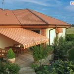 Zdjęcie prezentuje dom jednorodzinny o jasnej, kremowej elewacji z zainstalowaną markizą Mezzo mol, która osłania drewniany taras. Markiza charakteryzuje się poszyciem w ciepłej kolorystyce, składającym się z naprzemiennych pasów w odcieniach pomarańczu, czerwieni i beżu. Tkanina jest w pełni rozłożona pod kątem, wsparta na bocznych ramionach, a jej krawędź wykończona jest falistą falbaną z białą lamówką. Budynek posiada dach wielospadowy pokryty ceglastą dachówką ceramiczną, z wyraźnie zaznaczonymi brązowymi rynnami i podbitką dachową w kolorze jasnego drewna. Na tarasie pod markizą Mezzo mol znajduje się drewniany komplet mebli: prostokątny stół, krzesła z ażurowymi oparciami oraz ławka. Przestrzeń wokół tarasu jest bogato zagospodarowana roślinnością – widoczne są liczne krzewy ozdobne, iglaki oraz kwiaty w ceramicznych donicach. Po prawej stronie, nad mniejszym oknem, zamontowana jest dodatkowa, mniejsza markiza koszowa o identycznym wzorze materiału. W tle rozpościera się widok na zielony ogród z krętą ścieżką oraz daleki horyzont z linią lasu pod błękitnym, lekko zachmurzonym niebem.