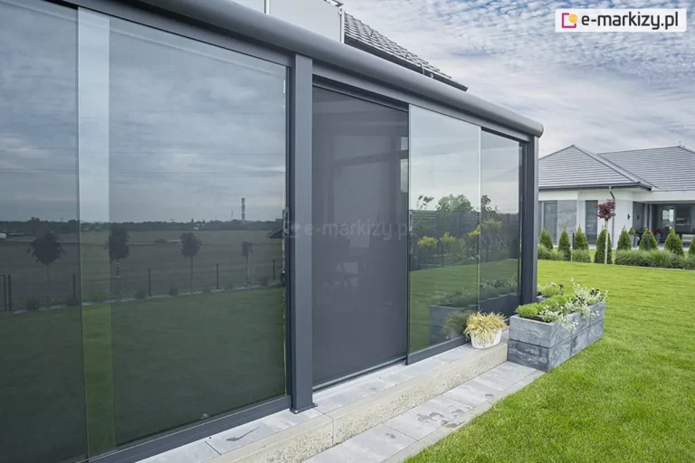 Nowoczesna, przeszklona konstrukcja ogrodu zimowego z zewnętrzną osłoną przeciwsłoneczną Refleksol, wkomponowaną w minimalistyczną zabudowę aluminiową w kolorze antracytowym. Refleksol wyposażony jest w prowadnice boczne i kasetę, a jego tkanina screen o wysokiej przezierności, w ciemnoszarym odcieniu, subtelnie zaciemnia wnętrze, jednocześnie umożliwiając widoczność na zewnątrz. System zamontowany na jednej z ruchomych tafli szklanych, osłaniających przestrzeń wypoczynkową pod dachem. W tle widoczna zadbana zieleń ogrodu, trawnik, rabaty z ozdobnymi roślinami oraz nowoczesny dom z płaskim dachem. W odbiciu na szybach widać panoramiczny widok na otwarte pola, drzewa i niebo, co dodaje przestrzeni lekkości. Całość stanowi przykład estetycznego i funkcjonalnego połączenia nowoczesnej architektury, technologii ochrony przeciwsłonecznej oraz natury.