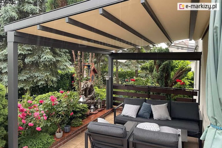 Widok z wnętrza tarasu na zadaszenie pergoli tkaninowej Square. Poszycie dachowe wykonane jest z jasnego materiału w odcieniu beżowym, który jest rozpięty na prowadnicach, tworząc płaską, lekko nachyloną powierzchnię. Konstrukcja pergoli wykonana została z aluminium, dach wsparty jest na dwóch solidnych podporach w kolorze antracytowym. Pod zadaszeniem, przy jasnej ścianie budynku, znajduje się strefa wypoczynkowa z nowoczesnymi meblami ogrodowymi: dużą, ciemnoszarą sofą z dekoracyjnymi poduszkami w paski i gładkimi szarymi oraz dwoma pasującymi fotelami ustawionymi przodem do siebie. Podłoga tarasu wyłożona jest ceramicznymi płytkami imitującymi jasne drewno. Taras otoczony jest niską balustradą z ciemnych, poziomych profili drewnianych lub kompozytowych, na której wiszą skrzynki z kwitnącymi czerwonymi pelargoniami. Przestrzeń wokół tarasu wypełnia bogata i gęsta roślinność ogrodowa – po lewej stronie widoczne są krzewy róż o intensywnie różowych kwiatach, a w tle liczne drzewa iglaste, w tym wysokie świerki srebrzyste. W głębi ogrodu, za pergolą, dostrzec można dekoracyjną rzeźbę ogrodową wykonaną z naturalnego kamienia lub pnia, obok której ustawiono lampiony oraz ozdobną lampę stojącą. Po prawej stronie kadru widoczny jest fragment pionowej zasłony tarasowej w kolorze jasnej zieleni, która jest spięta przy ścianie.