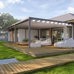 Zdjęcie przedstawia nowoczesną aranżację dużego tarasu wypoczynkowego z przyścienną pergolą aluminiową Tarasola Flat w kolorze antracytowym. Zadaszenie pergoli wykonane jest z białej tkaniny ułożonej w dekoracyjne, miękkie fale, co nadaje konstrukcji lekkości. Taras oraz prowadząca do niego ścieżka zostały zbudowane z ryflowanych desek drewnianych w naturalnym odcieniu. Na powierzchni tarasu umieszczono luksusowe meble ogrodowe o obłych kształtach z białej plecionki, w tym dwa duże szezlongi z jasnoszarymi poduchami oraz pasujący stolik kawowy. W głębi widoczne są również drewniane leżaki. Budynek o białej elewacji posiada liczne przeszklenia oraz drewniane detale wykończeniowe, które harmonizują z otaczającą go zielenią. Całość znajduje się na tle zadbanego trawnika oraz gęstego szpaleru drzew, w pełnym świetle dziennym przy lekko zachmurzonym niebie.