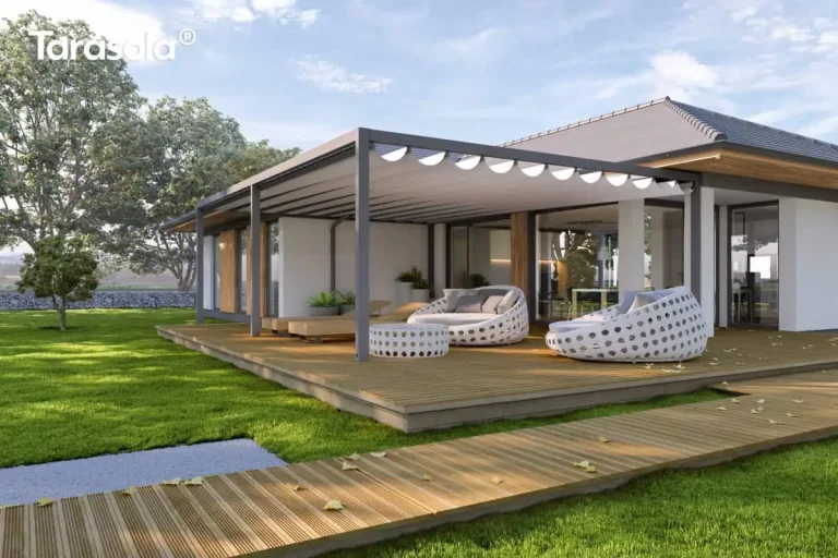 Zdjęcie przedstawia nowoczesną aranżację dużego tarasu wypoczynkowego z przyścienną pergolą aluminiową Tarasola Flat w kolorze antracytowym. Zadaszenie pergoli wykonane jest z białej tkaniny ułożonej w dekoracyjne, miękkie fale, co nadaje konstrukcji lekkości. Taras oraz prowadząca do niego ścieżka zostały zbudowane z ryflowanych desek drewnianych w naturalnym odcieniu. Na powierzchni tarasu umieszczono luksusowe meble ogrodowe o obłych kształtach z białej plecionki, w tym dwa duże szezlongi z jasnoszarymi poduchami oraz pasujący stolik kawowy. W głębi widoczne są również drewniane leżaki. Budynek o białej elewacji posiada liczne przeszklenia oraz drewniane detale wykończeniowe, które harmonizują z otaczającą go zielenią. Całość znajduje się na tle zadbanego trawnika oraz gęstego szpaleru drzew, w pełnym świetle dziennym przy lekko zachmurzonym niebie.