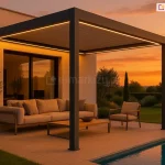 Nowoczesna pergola lamelowa Marlo w kolorze antracytowym, zainstalowana na przestronnym tarasie przy basenie w porze zachodu słońca. Konstrukcja jest zamontowana jedną stroną do jasnej elewacji budynku, a z przodu wspiera się na dwóch solidnych, kwadratowych profilach aluminiowych. Charakterystycznym elementem tej realizacji jest zintegrowane oświetlenie LED w formie liniowych pasów biegnących wzdłuż wewnętrznej krawędzi ramy zadaszenia, które emituje ciepłe, żółte światło. Pod pergolą zaaranżowano strefę wypoczynkową z beżową sofą ogrodową, fotelem oraz drewnianym stolikiem kawowym, ustawionymi na jasnych płytach tarasowych. Po lewej stronie widoczna jest roślina w donicy, a w tle duże przeszklenia prowadzące do wnętrza domu. Po prawej stronie kadru znajduje się fragment niecki basenowej z błękitną wodą, a dalej rozpościera się widok na ogród z przystrzyżonymi krzewami i drzewami na tle intensywnie pomarańczowego nieba. Lamele dachowe są całkowicie zamknięte, tworząc szczelne zadaszenie nad meblami ogrodowymi. Całość utrzymana w pomarańczowo-żółtym tonie zachodzącego słońca.