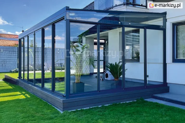 Zdjęcie przedstawia nowoczesną, przeszkloną zabudowę tarasu o konstrukcji aluminiowej w kolorze ciemnego antracytu, połączoną z bryłą białego, piętrowego budynku mieszkalnego. System przesuwnych drzwi szklanych Sotro tworzy pełne oszklenie ścian bocznych i frontowych, zapewniając transparentność i ochronę wnętrza, w którym widoczny jest ciemny, drewniany taras, rośliny egzotyczne w donicach oraz mała buda dla psa. Zadaszenie konstrukcji jest lekko nachylone i wykonane z przezroczystych paneli, a górne naświetla boczne mają trójkątny kształt dopasowany do skosu dachu i są mocowane punktowo za pomocą metalowych uchwytów. Całość posadowiona jest na podwyższonym fundamencie wykończonym deską kompozytową, który sąsiaduje z zadbanym trawnikiem, tworząc spójną strefę rekreacyjną zintegrowaną z ogrodem.