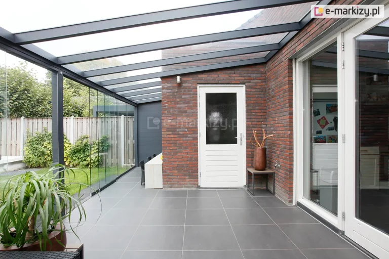 Nowoczesna realizacja zabudowy tarasu zintegrowana z bryłą budynku z czerwonej cegły. Konstrukcja zadaszenia Terano została wykonana z profili aluminiowych w kolorze antracytowym, które tworzą ramę dachu zamontowaną jednostronnie do elewacji budynku. Dach jest w całości przeszklony, podzielony na kilka podłużnych sekcji, co zapewnia pełną przezierność i ochronę przed opadami. Ściana boczna tarasu została zabudowana bezramowymi panelami szklanymi, które umożliwiają swobodny widok na ogród. Podłoże tarasu wyłożono wielkoformatowymi płytami gresowymi w ciemnoszarym odcieniu o matowym wykończeniu. Na tarasie, przy ceglanej ścianie, znajduje się biały stół jadalniany z ciemnymi krzesłami, a obok białych drzwi wejściowych ustawiono dekoracyjną miedzianą wazę na niskim stoliku. W narożniku na pierwszym planie widoczna jest duża roślina doniczkowa o długich, zielonych liściach. W tle, za szklaną ścianą, znajduje się drewniane ogrodzenie oraz gęsta roślinność ogrodowa, co tworzy prywatną i estetyczną przestrzeń do wypoczynku.