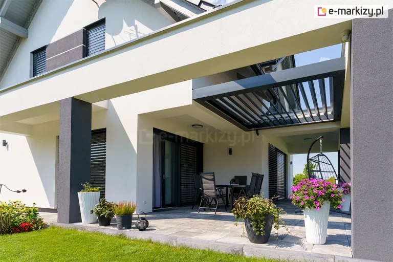 Zdjęcie prezentuje nowoczesny budynek mieszkalny o białej elewacji z zainstalowanym systemem dachu lamelowego SB400R. Konstrukcja zadaszenia jest wkomponowana w bryłę tarasu wspartą na masywnych, ciemnoszarych kolumnach i białych belkach konstrukcyjnych. Aluminiowe lamele w kolorze antracytowym znajdują się w pozycji uchylonej, co pozwala na regulację wpadającego światła słonecznego do strefy wypoczynkowej. Pod zadaszeniem widoczny jest stół z czarnymi krzesłami oraz wiszący fotel ogrodowy typu kokon z szarymi poduszkami. Taras wyłożony jest dużymi, jasnoszarymi płytami betonowymi, na których ustawiono liczne donice z kwitnącymi roślinami, w tym różowymi petuniami oraz wrzosami. W tle widoczne są nowoczesne żaluzje fasadowe w kolorze antracytowym, osłaniające duże przeszklenia budynku, a po lewej stronie znajduje się fragment zadbanego trawnika. Całość przedstawiona jest w pełnym świetle dziennym, co podkreśla kontrastową kolorystykę materiałów i nowoczesną architekturę obiektu.