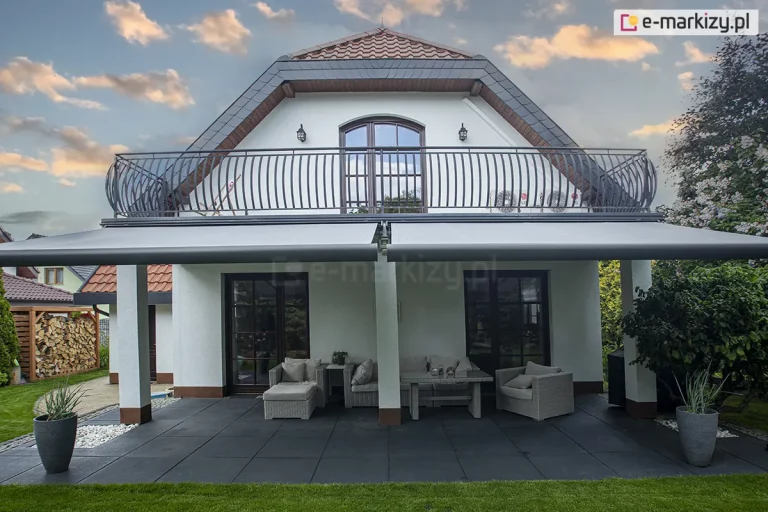 Zdjęcie przedstawia szeroki widok na tylną elewację jednorodzinnego domu z poddaszem użytkowym oraz przestronnym tarasem parterowym, który został osłonięty przez dwie zestawione obok siebie markizy tarasowe Palladio Selt. Konstrukcja markiz wykonana została w kolorze srebrzysty grafit a poszycie wykonano z jasnoszarej tkaniny akrylowej. Budynek posiada białą elewację, a jego bryłę wieńczy dwuspadowy dach o łamanej konstrukcji, pokryty dachówką w kolorze ceglastym z ciemnoszarym wykończeniem krawędzi. Na piętrze znajduje się balkon z ozdobną, zaokrągloną balustradą z kutej stali w ciemnym kolorze, rozciągający się na całą szerokość elewacji. Pod balkonem zamontowano system dwóch markiz z napędem elektrycznym, których ramiona są w pełni rozłożone, tworząc głęboki cień nad strefą wypoczynkową. Taras został wyłożony wielkoformatowymi płytami w kolorze grafitowym. Na tarasie ustawiono zestaw mebli ogrodowych wykonanych z szarego technorattanu, składający się z narożnej sofy, fotela oraz prostokątnego stolika kawowego, z dopasowanymi kolorystycznie poduszkami. Po bokach tarasu znajdują się symetrycznie ustawione, wysokie donice w kolorze ciemnoszarym z ozdobnymi trawami. W tle, po lewej stronie, widoczna jest drewniana wiata na drewno kominkowe, a po prawej gęsta zieleń ogrodowa. Niebo nad domem jest lekko zachmurzone z widocznymi promieniami zachodzącego słońca, co nadaje scenie ciepłą kolorystykę.