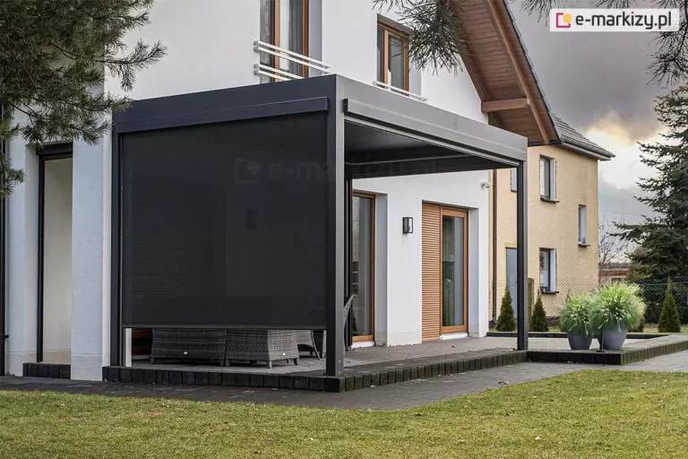 Nowoczesna pergola tarasowa w kolorze antracytowym, zamontowana przy białej elewacji piętrowego domu jednorodzinnego, ukazana w świetle dziennym przy pochmurnym niebie. Konstrukcja posiada w pełni opuszczoną, ciemną roletę boczną typu screen po lewej stronie, która niemal całkowicie przesłania wnętrze tarasu. Frontowa część pergoli pozostaje otwarta, odsłaniając fragmenty szarych mebli ogrodowych z technorattanowymi plecionkami. Na elewacji budynku widoczne są brązowe ramy okienne, białe barierki na piętrze oraz pionowa roleta zewnętrzna w kolorze drewna na parterze. Taras wykonany jest z ciemnej kostki brukowej, a przed nim znajduje się zadbany trawnik. Po prawej stronie, na obrzeżu tarasu, stoją dwie szare donice z kępami ozdobnych traw, a w tle widoczny jest sąsiedni dom o żółtej elewacji oraz iglaki.