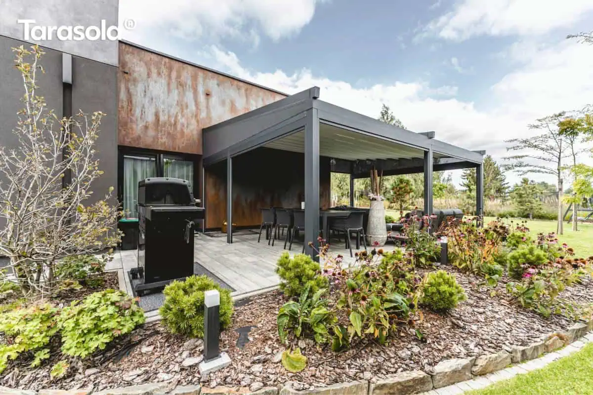 Nowoczesna zabudowa tarasu z wykorzystaniem przyściennej pergoli Elegancy Tarasola w kolorze antracytowym, zintegrowanej z bryłą budynku o elewacji imitującej rdzawą stal corten. Konstrukcja posiada rozsuwany dach tkaninowy w jasnym kolorze, który w tej konfiguracji pozostaje częściowo osłonięty bocznymi roletami screen, zapewniającymi prywatność i ochronę przed słońcem. Na utwardzonym jasną kostką tarasie znajduje się duży stół obiadowy z krzesłami oraz nowoczesny czarny grill gazowy ustawiony z boku. Przed tarasem zaprojektowano estetyczne rabaty ogrodowe wysypane korą, obsadzone ozdobnymi krzewami, iglakami oraz kwitnącymi bylinami, a cała strefa wypoczynkowa płynnie łączy się z rozległym trawnikiem w głębi ogrodu.