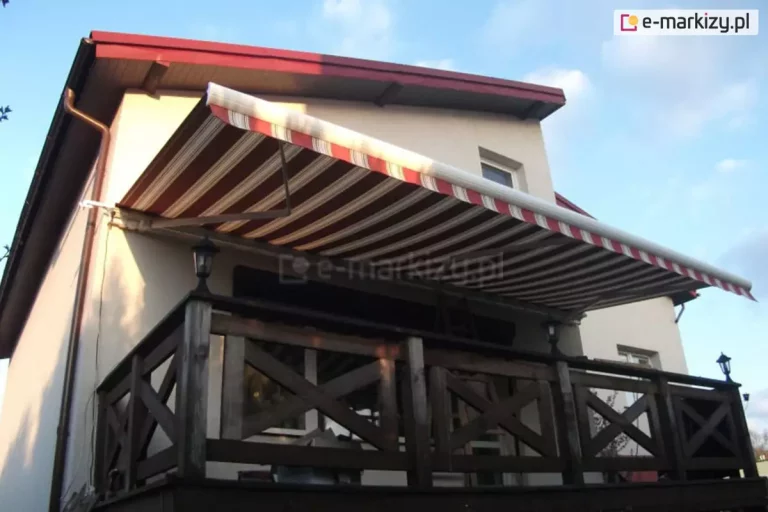 Zdjęcie prezentuje szeroką markizę tarasową Palladio Selt zamontowaną na elewacji piętrowego domu jednorodzinnego, osłaniającą przestronny balkon. Poszycie markizy wykonane jest z materiału akrylowego w klasyczne, poziome pasy w kolorystyce bordowej, brązowej i beżowej, co harmonizuje z ciemną stolarką oraz dachem budynku. Markiza jest w pełni rozwinięta, wsparta na białych ramionach przegubowych zamontowanych do jasnej, tynkowanej ściany. Balkon zabezpieczony jest solidną, drewnianą barierką o ciemnobrązowym odcieniu, charakteryzującą się konstrukcją z krzyżującymi się belkami wypełniającymi poszczególne przęsła. Na balustradzie zamontowane są klasyczne, czarne lampiony zewnętrzne pełniące funkcję oświetlenia. Budynek posiada dwuspadowy dach o ciemnoczerwonym pokryciu z widocznymi podbitkami dachowymi w kolorze naturalnego drewna. Po lewej stronie fasady widoczna jest miedziana rura spustowa odprowadzająca wodę z rynny. Całość kompozycji uchwycona jest z dolnej perspektywy na tle błękitnego nieba z delikatnymi chmurami, co podkreśla funkcjonalność osłony przeciwsłonecznej w słoneczny dzień.
