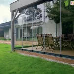 Nowoczesna zabudowa tarasu wykonana z systemu bezramowych przesuwnych szyb, zamontowana przy jednokondygnacyjnym budynku mieszkalnym. Konstrukcja oparta na smukłych, aluminiowych profilach, z przezroczystymi panelami szklanymi Miros osadzonymi w dolnej i górnej prowadnicy. Wewnątrz znajduje się drewniany zestaw mebli ogrodowych – duży stół oraz kilka krzeseł składanych. Otoczenie to zadbany trawnik, rabaty z białym grysem i taras z drewnianych desek. Przeszklenie odbija sylwetki drzew i fragmenty otoczenia.