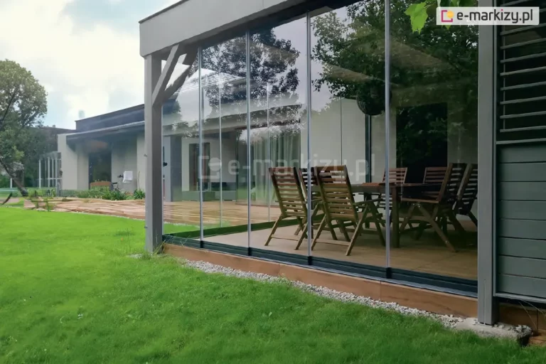 Nowoczesna zabudowa tarasu wykonana z systemu bezramowych przesuwnych szyb, zamontowana przy jednokondygnacyjnym budynku mieszkalnym. Konstrukcja oparta na smukłych, aluminiowych profilach, z przezroczystymi panelami szklanymi Miros osadzonymi w dolnej i górnej prowadnicy. Wewnątrz znajduje się drewniany zestaw mebli ogrodowych – duży stół oraz kilka krzeseł składanych. Otoczenie to zadbany trawnik, rabaty z białym grysem i taras z drewnianych desek. Przeszklenie odbija sylwetki drzew i fragmenty otoczenia.