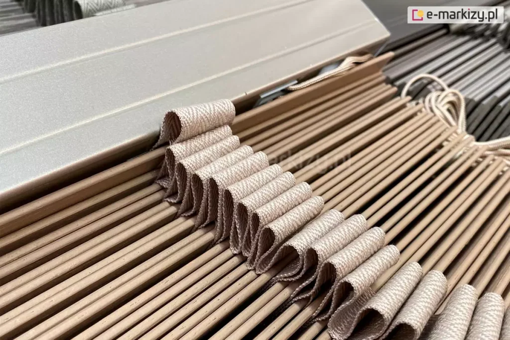 Żaluzja drewniana z lamelami 5 cm w kolorze naturalnego drewna z wyraźnie widoczną ułożoną w fale drabinką taśmową dopasowaną do kolorystyki lamel. Powyżej pakietu lameli widoczna rynna górna aluminiowa, także wykonana w kolorze zbliżonym do barwy lamel żaluzji.