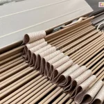 Żaluzja drewniana z lamelami 5 cm w kolorze naturalnego drewna z wyraźnie widoczną ułożoną w fale drabinką taśmową dopasowaną do kolorystyki lamel. Powyżej pakietu lameli widoczna rynna górna aluminiowa, także wykonana w kolorze zbliżonym do barwy lamel żaluzji.
