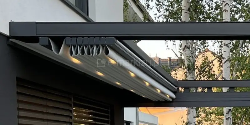 Zbliżenie dachu pergoli Square Flat w pozycji zwiniętej. Pomiędzy faldami jasnej tkaniny widoczne jest ciepłe światło zamocowanych w belkach przeciwwietrznych lampek LED. Dach przymocowany do zawieszonej na ścianie aluminiowej konstrukcji w kolorze grafitowym. W tle widoczne brzozy oraz fragment sąsiedniego budynku o żółtej elewacji.