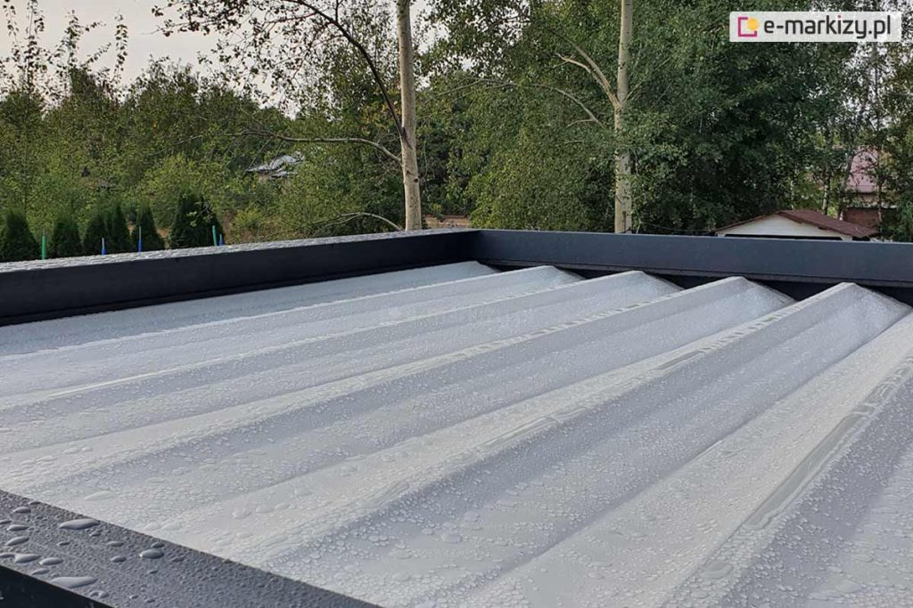 Zdjęcie przedstawia widok z góry na rozłożone zadaszenie pergoli tarasowej Luxury wykonane z jasnej, wodoodpornej tkaniny w kolorze białym. Powierzchnia materiału jest ułożona w regularne, podłużne segmenty wsparte na poprzecznych profilach, a na jej powierzchni widoczne są liczne krople wody, co demonstruje właściwości hydrofobowe tkaniny. Rama pergoli otaczająca poszycie wykonana jest z szerokich profili aluminiowych w kolorze ciemnego antracytu o matowym wykończeniu. W tle, poza konstrukcją, widoczna jest gęsta roślinność ogrodowa z przewagą drzew liściastych o zielonych i lekko żółknących liściach oraz zarys dachu sąsiedniego budynku. Perspektywa zdjęcia z bliska podkreśla skuteczność ochrony przed opadami atmosferycznymi oraz solidność wykonania systemu odprowadzania wody w nowoczesnej zabudowie tarasowej.