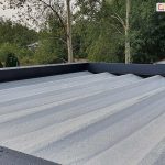 Zdjęcie przedstawia widok z góry na rozłożone zadaszenie pergoli tarasowej Luxury wykonane z jasnej, wodoodpornej tkaniny w kolorze białym. Powierzchnia materiału jest ułożona w regularne, podłużne segmenty wsparte na poprzecznych profilach, a na jej powierzchni widoczne są liczne krople wody, co demonstruje właściwości hydrofobowe tkaniny. Rama pergoli otaczająca poszycie wykonana jest z szerokich profili aluminiowych w kolorze ciemnego antracytu o matowym wykończeniu. W tle, poza konstrukcją, widoczna jest gęsta roślinność ogrodowa z przewagą drzew liściastych o zielonych i lekko żółknących liściach oraz zarys dachu sąsiedniego budynku. Perspektywa zdjęcia z bliska podkreśla skuteczność ochrony przed opadami atmosferycznymi oraz solidność wykonania systemu odprowadzania wody w nowoczesnej zabudowie tarasowej.