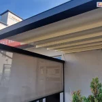 Zdjęcie prezentuje zbliżenie na górną część konstrukcji pergoli tarasowej zamontowanej przy jasnoszarej elewacji budynku. Głównym elementem jest pozioma belka konstrukcyjna w kolorze ciemnego antracytu. Po lewej stronie widoczna jest częściowo opuszczony refelksol ZIP wykonany z półtransparentnej tkaniny w odcieniu szarym, przez którą prześwitują kontury otoczenia. Powyżej rolety, pod głównym profilem, znajduje się rozłożone zadaszenie tkaninowe w kolorze kremowym, ułożone w regularne, poziome fałdy. Konstrukcja zadaszenia jest zintegrowana z aluminiowymi profilami poprzecznymi. W prawym dolnym rogu kadru widoczne jest gęste, zielone igliwie krzewu ozdobnego, a w tle zarysowuje się biała bryła sąsiedniego budynku z widocznym fragmentem napisu na czerwonym tle. Całość ujęcia została wykonana w świetle dziennym, które podkreśla fakturę materiałów oraz precyzję łączenia elementów metalowych z tekstylnymi.