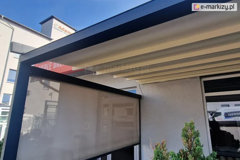 Zdjęcie prezentuje zbliżenie na górną część konstrukcji pergoli tarasowej zamontowanej przy jasnoszarej elewacji budynku. Głównym elementem jest pozioma belka konstrukcyjna w kolorze ciemnego antracytu. Po lewej stronie widoczna jest częściowo opuszczony refelksol ZIP wykonany z półtransparentnej tkaniny w odcieniu szarym, przez którą prześwitują kontury otoczenia. Powyżej rolety, pod głównym profilem, znajduje się rozłożone zadaszenie tkaninowe w kolorze kremowym, ułożone w regularne, poziome fałdy. Konstrukcja zadaszenia jest zintegrowana z aluminiowymi profilami poprzecznymi. W prawym dolnym rogu kadru widoczne jest gęste, zielone igliwie krzewu ozdobnego, a w tle zarysowuje się biała bryła sąsiedniego budynku z widocznym fragmentem napisu na czerwonym tle. Całość ujęcia została wykonana w świetle dziennym, które podkreśla fakturę materiałów oraz precyzję łączenia elementów metalowych z tekstylnymi.