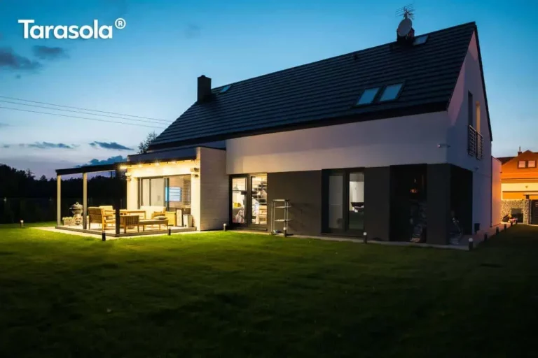 Nowoczesna, dwukolorowa bryła budynku mieszkalnego z poddaszem użytkowym i dachem dwuspadowym pokrytym ciemną dachówką, sfotografowana w scenerii wieczornej z włączonym oświetleniem zewnętrznym. Do bocznej elewacji domu przylega aluminiowa pergola tarasowa w kolorze antracytowym z ruchomym, tkaninowym zadaszeniem w jasnym odcieniu. Konstrukcja pergoli opiera się na solidnych profilach pionowych, a pod zadaszeniem znajduje się strefa wypoczynkowa wyposażona w drewniane meble ogrodowe: sofę, fotele oraz stolik kawowy. Oświetlenie tarasu realizowane jest za pomocą punktowych reflektorów zamontowanych na elewacji oraz zintegrowanych listew LED w konstrukcji zadaszenia, co tworzy ciepłą poświatę na jasnej ścianie budynku. Teren wokół domu pokrywa zadbany trawnik, na którym rozmieszczono niskie słupki oświetleniowe wyznaczające linię ogrodu. Elewacja parteru charakteryzuje się dużymi przeszkleniami i ciemnym tynkiem w dolnej partii, który kontrastuje z białą elewacją piętra.