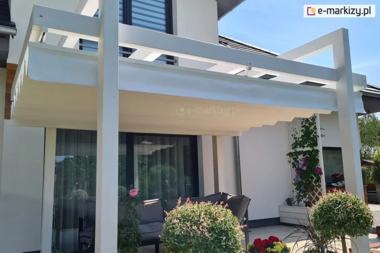 Pergola drewniana kolumnowa Moza z opcjonalnym dachem rzymskim Supro w odcieniach bieli doskonale współgra z białą fasadą budynku oraz pełnym roślinności otoczeniem tarasu.