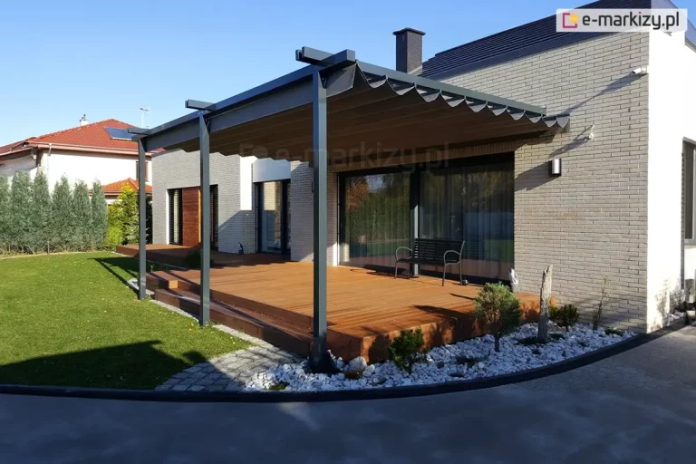 Zdjęcie prezentuje nowoczesną, przyścienną pergolę o konstrukcji aluminiowej w kolorze antracytowym, zamontowaną nad rozległym tarasem wykonanym z naturalnych desek drewnianych. Na pergoli zastosowany został system zadaszenia Supro, z tkaniną w kolorze ciemnoszarym (antracytowym), która jest niemal całkowicie rozwinięta, tworząc regularne, płytkie fale podwieszone pod poprzecznymi profilami. Konstrukcja wspiera się na trzech pionowych słupach o kwadratowym przekroju, zakotwiczonych w podłożu otoczonym dekoracyjną opaską z białych kamieni i niskimi nasadzeniami iglaków. Budynek, do którego przylega pergola, ma nowoczesną bryłę wykończoną jasnoszarą cegłą elewacyjną oraz duże, przesuwne przeszklenia tarasowe, przed którymi stoi minimalistyczna czarna ławka. W tle widoczny jest fragment sąsiedniej posesji z domem o czerwonym dachu oraz wysoki żywopłot z tui, a całość kompozycji oświetla ostre, popołudniowe słońce, rzucając wyraźne cienie na drewnianą nawierzchnię tarasu.