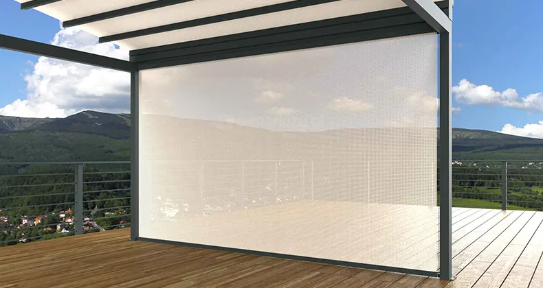 Pergola z dachem tkaninowym ustawiona na tarasie z drewna z metalową barierką. Frontowa ściana pergoli pomiędzy słupami osłonięta została roletą screen ZIP U dopasowaną kolorystycznie do pergoli, zastosowano w niej przezierną tkaninę, przez którą widać otoczenie tarasu, liczne zielone wzgórza, lasy oraz niebo z kilkoma chmurami.