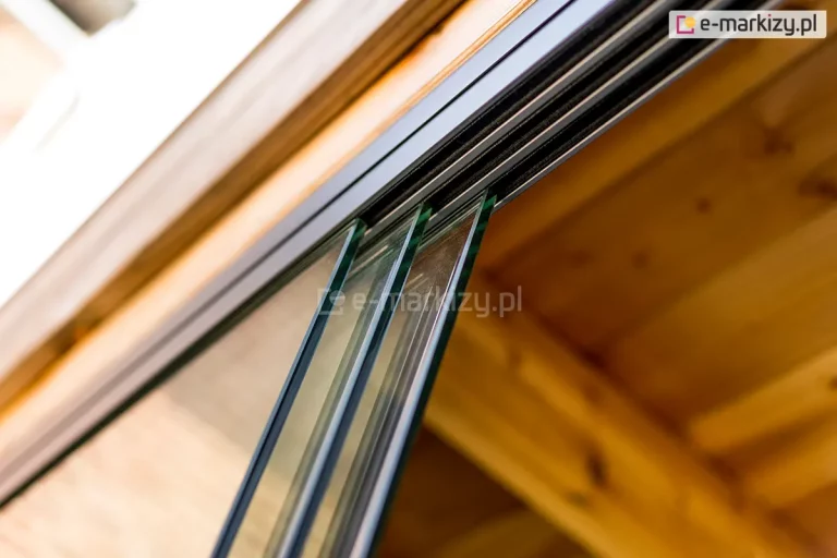 Zbliżenie na górną część systemu przesuwnych paneli szklanych (model Auris) zamontowanych na aluminiowej prowadnicy Fiano marki Deponti. Na pierwszym planie widoczne są trzy tafle hartowanego szkła o oszlifowanych krawędziach, które są ustawione równolegle względem siebie w wielotorowej szynie. Prowadnica o ciemnym, matowym wykończeniu w kolorze antracytowym jest przymocowana do drewnianej konstrukcji zadaszenia wykonanej z jasnych, sosnowych belek. Wewnątrz szyny widoczne są uszczelki szczotkowe, które zapewniają płynny ślizg paneli oraz ochronę przed wiatrem i zanieczyszczeniami. Perspektywa zdjęcia jest ujęta od dołu, pod kątem, co pozwala na dokładne przyjrzenie się mechanizmowi jezdnemu i grubości zastosowanego szkła.