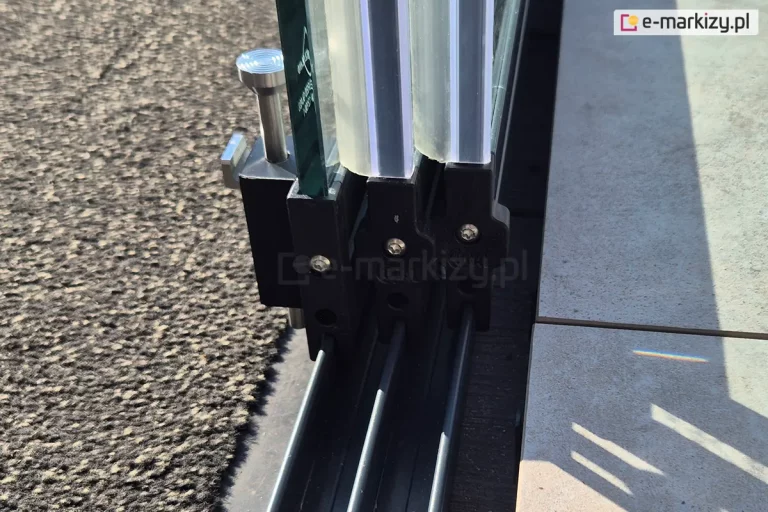 Prowadnica dolna z trzema panelami szklanymi, których krawędzie uzbrojono w listwy przeciwprzeciągowe gumowe, przejrzyste. Na ostatnim panelu, przy dywanie widać stoper, służący do zablokowania możliwości otwarcia drzwi z zewnątrz.