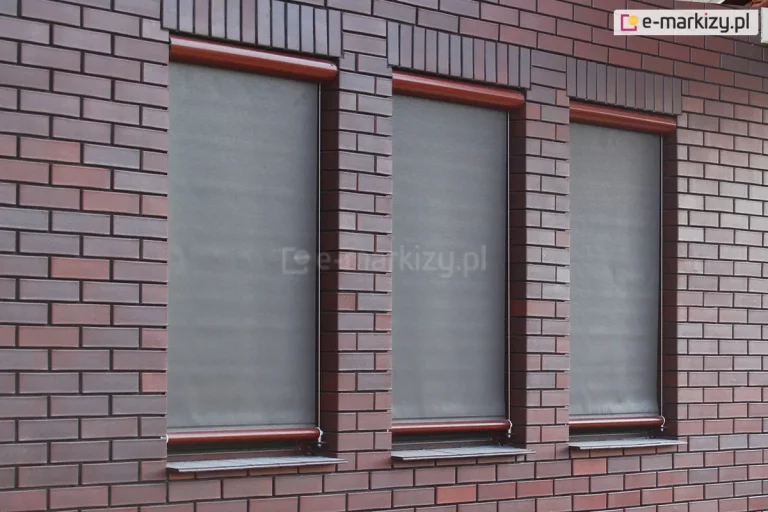 Elewacja z trzema dużymi, pionowymi oknami, na których zamontowano zewnętrzne rolety screenowe typu Refleksol 76. Każda osłona składa się z półtransparentnej tkaniny w kolorze szarym,zwijanej do aluminiowej kasety ukrytej w górnej części ramy. Kasety są lakierowane w kolorze ciemnego drewna, dopasowanego do otoczenia ceglanej elewacji. Z kasety do belki dolnej z tkaniną prowadzą boczne prowadnice linkowe zamocowane do ramy okna. Refleksol 76 to system osłon przeciwsłonecznych przeznaczony do montażu zewnętrznego, który łączy estetykę z funkcją ochrony przed nagrzewaniem. Tkanina zapewnia prywatność i rozproszenie światła, a jednocześnie nie zaciemnia wnętrza całkowicie. Widoczne na zdjęciu wykonanie świadczy o precyzji montażu i integracji systemu z architekturą budynku.