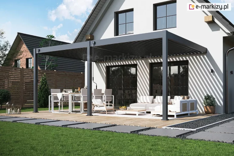 Pergola lamelowa przyścienna Rubio Soft w kolorze antracytowym, zainstalowana na przestronnym tarasie z jasnych desek, przylegająca do nowoczesnego, białego domu z dwuspadowym dachem. Konstrukcja zadaszenia opiera się na trzech solidnych, pionowych słupach o kwadratowym przekroju, z których środkowy stanowi punkt łączenia dwóch sekcji. Lamele dachu w obu modułach są ustawione pod kątem (półotwarte), co sprawia, że na jasnej elewacji budynku oraz na wielkoformatowych oknach tarasowych powstaje rytmiczny wzór ukośnych pasów światła i cienia. Pod lewym modułem pergoli znajduje się strefa jadalniana z dużym stołem i jasnymi krzesłami, natomiast pod prawym modułem zaaranżowano strefę relaksu z białą sofą narożną, fotelem oraz niskim stolikiem kawowym. Na belkach konstrukcyjnych widoczne są dwie czarne korby służące do ręcznej regulacji nachylenia lameli. Taras otoczony jest zadbanym, zielonym trawnikiem, przez który przebiega ścieżka z dużych, szarych płyt betonowych osadzonych w ciemnym kruszywie. W tle, za drewnianym ogrodzeniem, widoczny jest fragment sąsiedniego domu z ciemnym dachem, a nad całością rozciąga się pogodne niebo z delikatnymi chmurami.