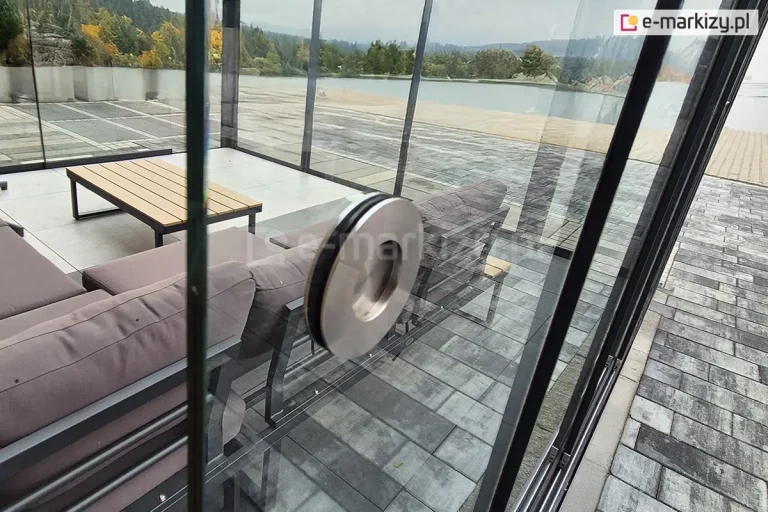 Zbliżenie srebrnego, metalowego uchwytu systemu Auris zamocowanego bezpośrednio do szklanego panelu przesuwnego. Tafla szkła jest przezroczysta, a uchwyt umieszczony jest na brzegu pośrodku wysokosci, umożliwiając wygodne przesuwanie panelu w prowadnicach. Konstrukcja uchwytu jest minimalistyczna, dopasowana wizualnie do systemu przesuwnych szyb tarasowych Auris. W obrębie zadaszenia widoczne dwa kolejne zsunięte panele szklane oraz sofa i stolik ustawione pod dachem ze szkła. W tle widoczna rzeka z piaszczystą plażą oraz wyłożonym kostka brukową zejściem do jej brzegu. Doskonale widoczne także drzewa porastające przeciwległy brzeg oraz lekko zachmurzone niebo nad nimi.