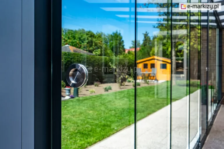 Zbliżenie na okrągły, metalowy uchwyt wpuszczany (muszlowy) zamontowany na tafli bezramowego szkła przesuwnego systemu Auris. Uchwyt wykonany ze stali nierdzewnej szczotkowanej jest zainstalowany w fabrycznym nawiercie szyby, co zapewnia stabilność i ergonomię podczas przesuwania paneli. Na szklanej powierzchni widoczne jest odbicie błękitnego nieba oraz zieleni ogrodowej. W tle, za przezroczystym przeszkleniem, rozpościera się widok na nasłoneczniony trawnik, roślinność ozdobną oraz drewniany domek narzędziowy w kolorze miodowym. Ostrość zdjęcia skoncentrowana jest na detalu okucia, podkreślając precyzję wykończenia krawędzi szkła hartowanego.